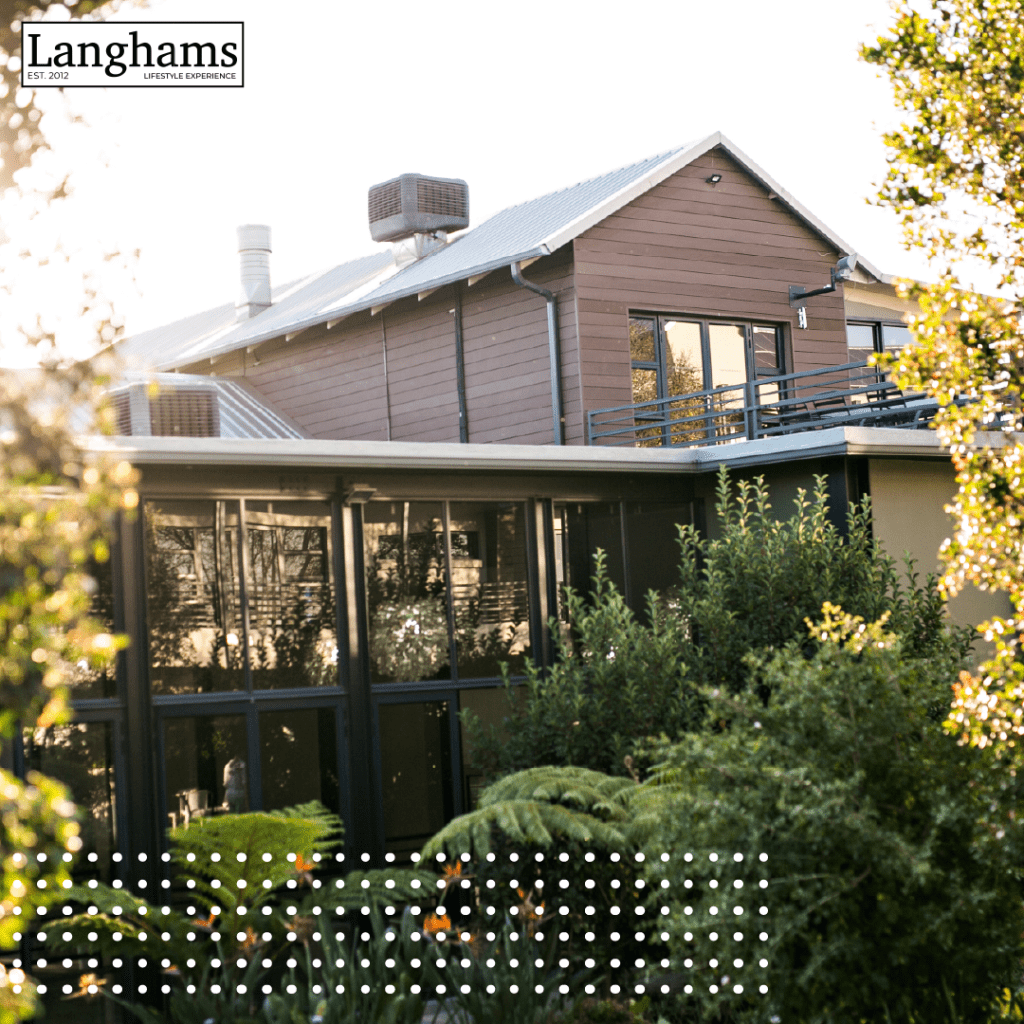 Exterior view of Langhams Lifestyle Experience, featuring modern glass-fronted architecture surrounded by lush greenery and natural sunlight.